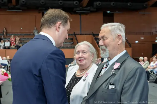 Spotkanie trzech osób, w tym mężczyzny w garniturze i starszej pary, w sali pełnej ludzi.