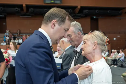Wręczenie odznaczenia przez mężczyznę starszej kobiecie podczas uroczystości.