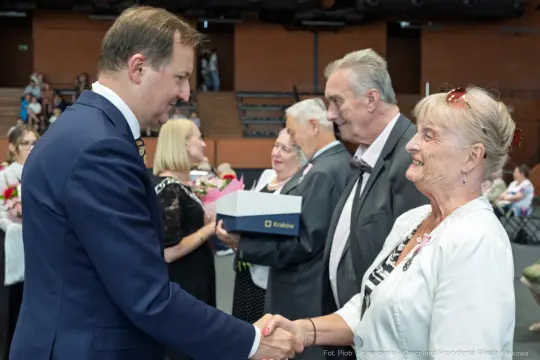 Mężczyzna w garniturze wita się z uśmiechniętą kobietą w białej bluzce. W tle inni uczestnicy wydarzenia.