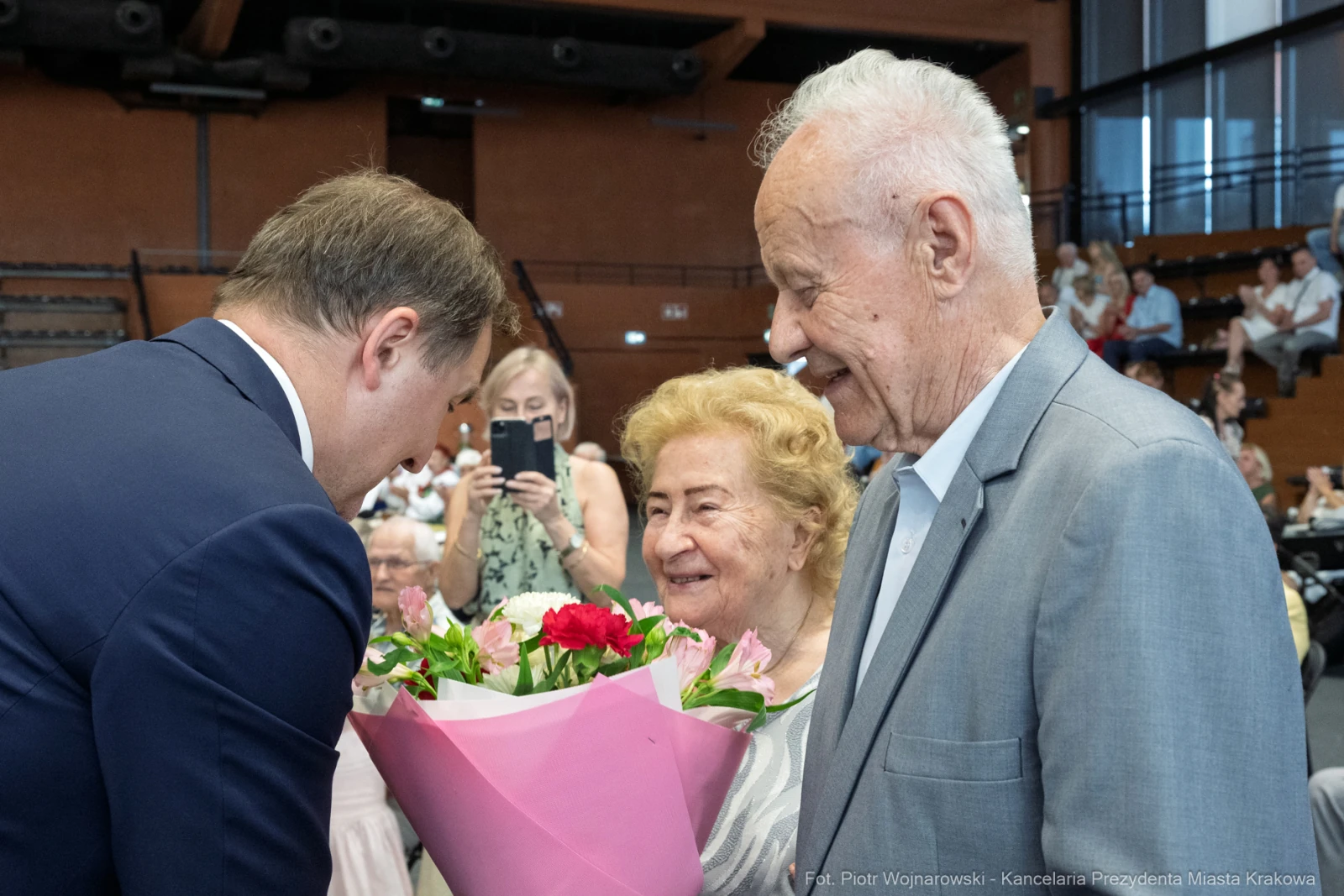 Spotkanie dwóch starszych osób z mężczyzną wręczającym bukiet kwiatów.  Autor: Piotr Wojnarowski