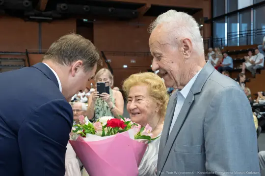 Spotkanie dwóch starszych osób z mężczyzną wręczającym bukiet kwiatów.