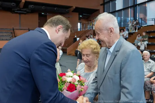 Mężczyzna wręcza bukiet kwiatów starszemu mężczyźnie, obok stoi starsza kobieta.