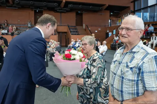 Mężczyzna wręcza bukiet kwiatów starszej kobiecie, obok stoi starszy mężczyzna. W tle zgromadzenie ludzi.