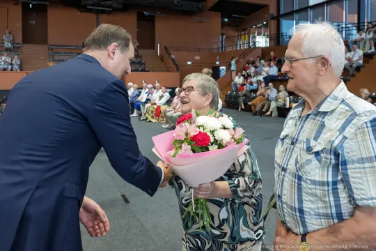 Mężczyzna wręcza bukiet kwiatów starszej kobiecie, obok stoi starszy mężczyzna. W tle widownia.