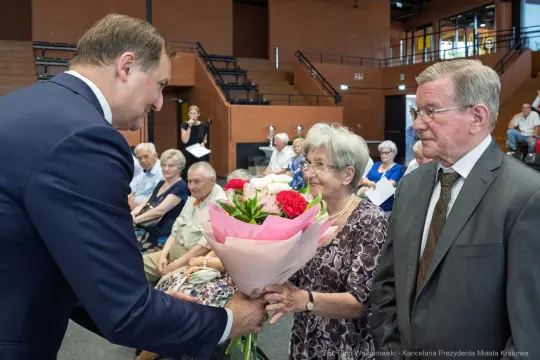 Mężczyzna wręcza bukiet kwiatów starszej kobiecie w towarzystwie innych osób.