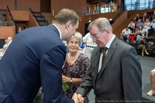 Mężczyzna w garniturze podaje rękę innemu mężczyźnie, w tle siedzi kobieta.