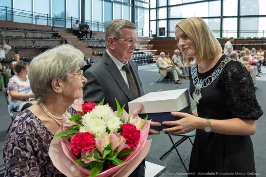 Kobieta wręcza prezent starszym osobom, w tle widoczna sala z siedzeniami.