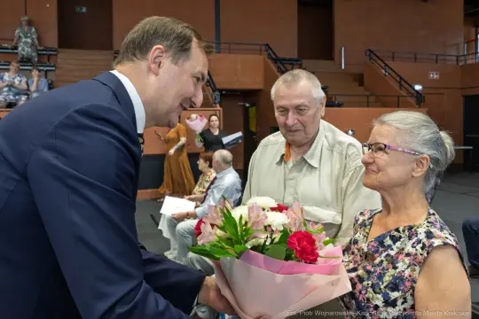 Mężczyzna wręcza bukiet kwiatów starszej kobiecie, obok stoi mężczyzna. W tle inni uczestnicy wydarzenia.