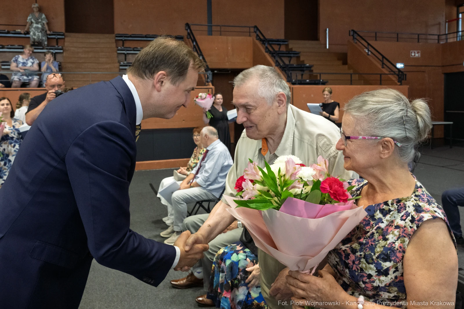 Mężczyzna w garniturze podaje rękę starszemu mężczyźnie, który trzyma bukiet kwiatów.  Autor: Piotr Wojnarowski