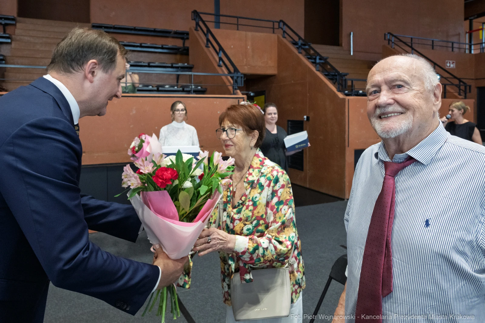 Mężczyzna wręcza bukiet kwiatów starszej parze w sali wydarzeń.  Autor: Piotr Wojnarowski