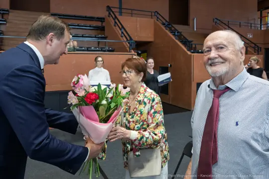 Mężczyzna wręcza bukiet kwiatów starszej parze w sali wydarzeń.