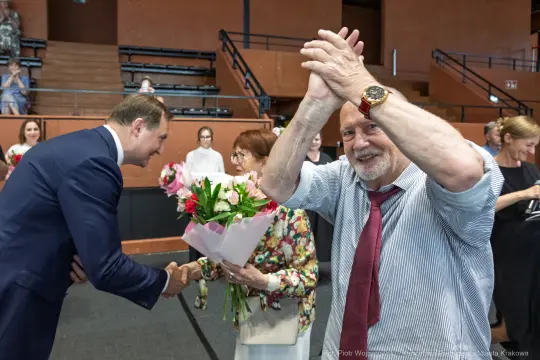 Mężczyzna w koszuli z uśmiechem klaska w dłonie, obok kobieta z bukietem kwiatów.