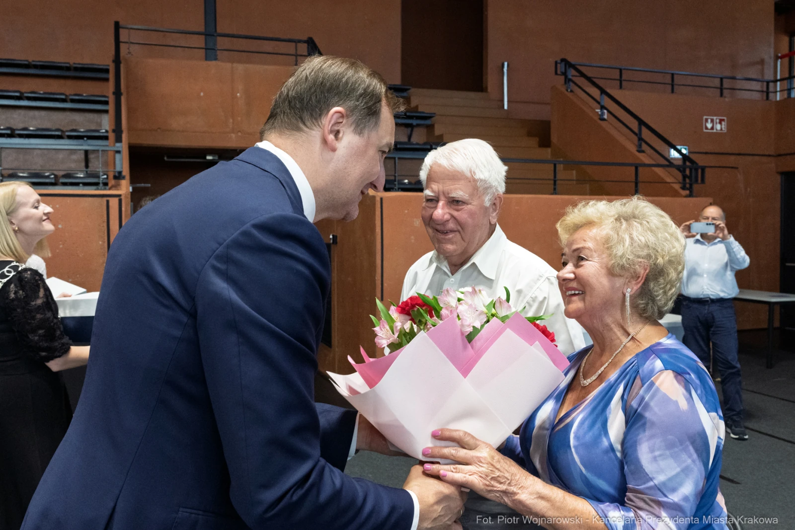 Mężczyzna wręcza bukiet kwiatów starszej kobiecie, obok stoi uśmiechający się mężczyzna.  Autor: Piotr Wojnarowski