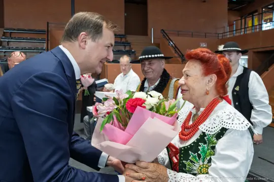 Mężczyzna wręcza bukiet kwiatów starszej kobiecie w tradycyjnym stroju ludowym.