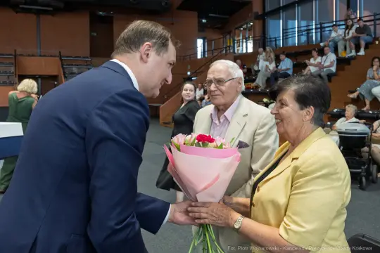 Mężczyzna wręcza bukiet kwiatów starszej kobiecie, obok stoi mężczyzna w okularach.