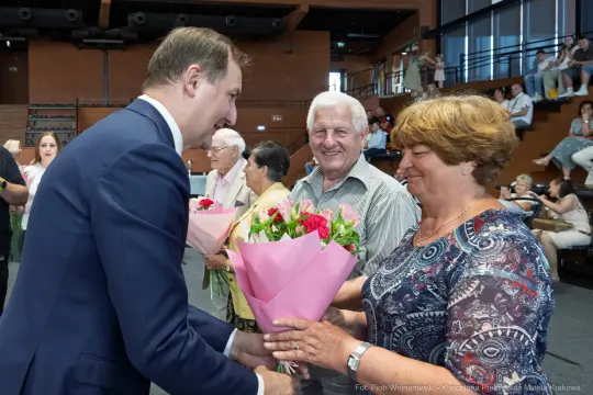 Mężczyzna wręcza bukiet kwiatów uśmiechniętej kobiecie w towarzystwie innych osób.