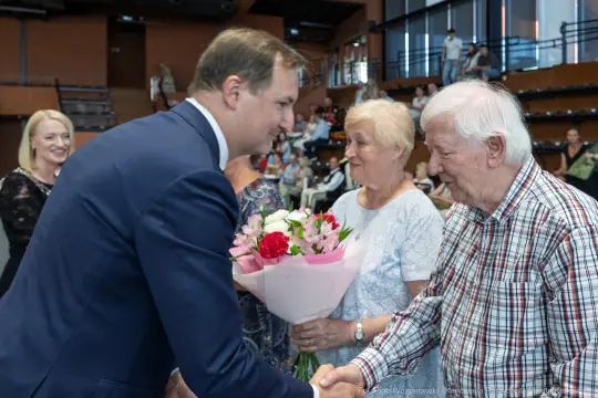 Mężczyzna w garniturze wita starsze małżeństwo, kobieta trzyma bukiet kwiatów.