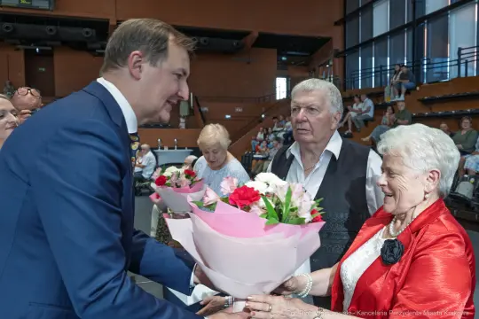 Mężczyzna wręcza kwiaty starszym osobom w sali.