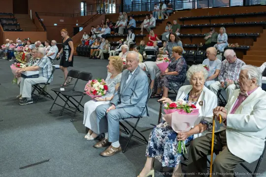 Grupa seniorów siedzących na widowni z bukietami kwiatów.