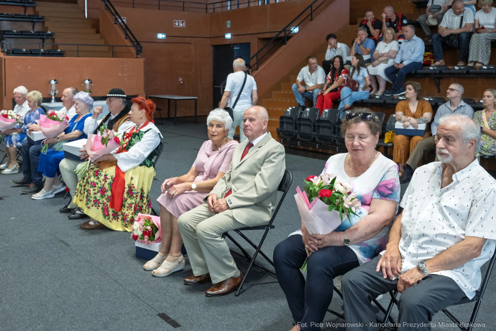 Grupa osób siedzących na widowni, trzymających kwiaty i uśmiechających się.  Autor: Piotr Wojnarowski