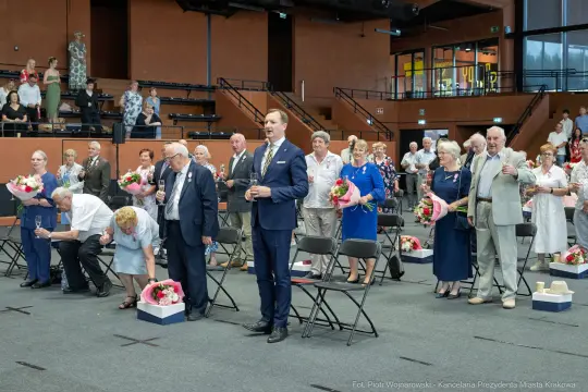 Uroczystość z udziałem osób starszych, kwiaty, stojący mężczyzna w garniturze.