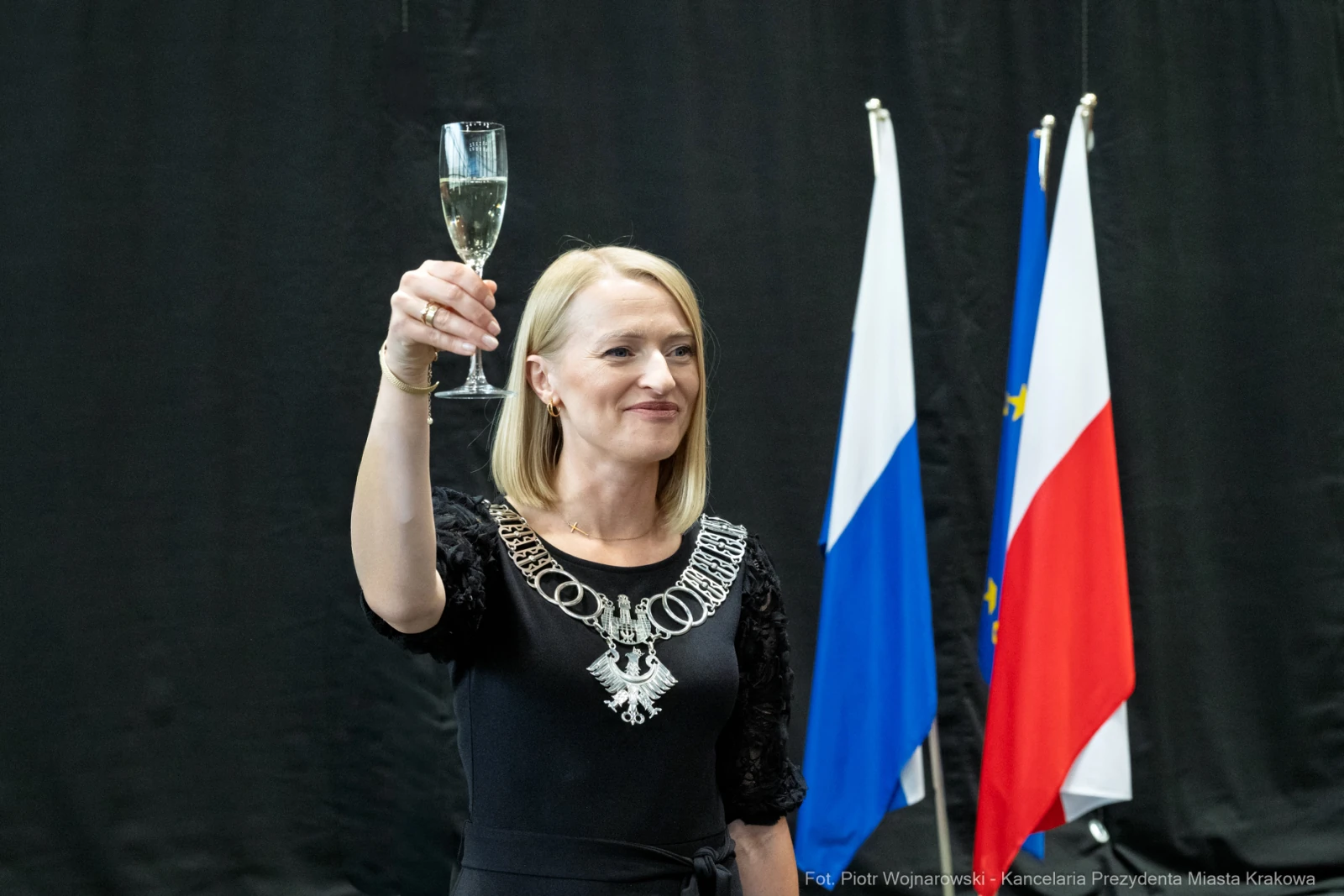 Kobieta w czarnej sukience wznosi toast z kieliszkiem szampana. W tle flagi Polski i Unii Europejskiej.  Autor: Piotr Wojnarowski