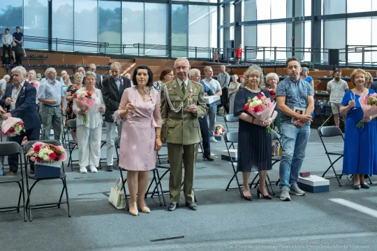Uroczystość z udziałem osób w eleganckich strojach, trzymających kwiaty i nagrody.