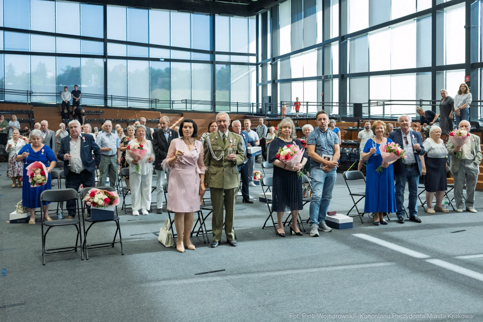 Grupa ludzi w sali, trzymających kwiaty, podczas uroczystości.  Autor: Piotr Wojnarowski