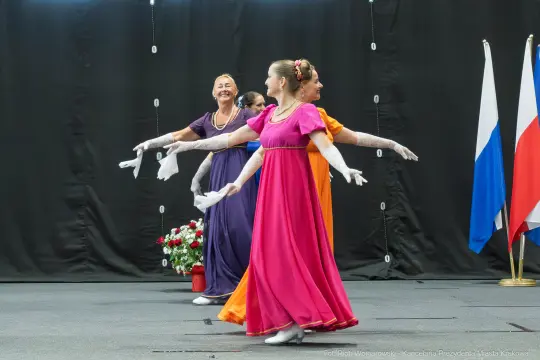 Cztery kobiety w eleganckich sukienkach tańczą na scenie, w tle flagi i kwiaty.
