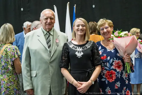 Trzy osoby uśmiechają się, jedna trzyma bukiet kwiatów, w tle flagi.