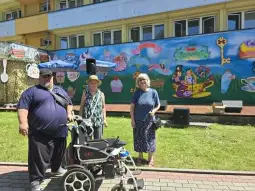 Trzy osoby stoją obok wózka inwalidzkiego przed kolorowym muralem.