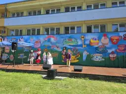 Dzieci w strojach występują na scenie przed kolorowym muralem z motywami kulinarnymi.