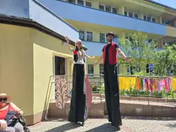 Dwie osoby na wysokich szczudłach w kolorowych strojach, obok siedząca kobieta w kapeluszu.