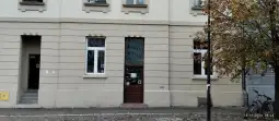 Budynek z jasną elewacją, drzwi wejściowe w centralnej części, obok okna i drzewo.