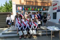 Grupa uczniów w strojach galowych z dyplomami na schodach szkoły.