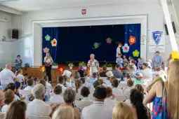 Uroczystość szkolna z dziećmi i nauczycielami na scenie, kolorowe dekoracje w tle.