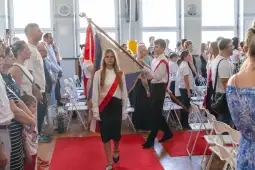 Uczniowie w białych koszulach niosą sztandar podczas ceremonii w sali.