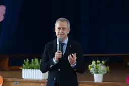 Mężczyzna w garniturze przemawia do publiczności, trzymając mikrofon. W tle dekoracje i rośliny.