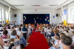 Uroczystość szkolna z mówcą na scenie, publiczność siedząca na krzesłach, dekoracje kwiatowe.