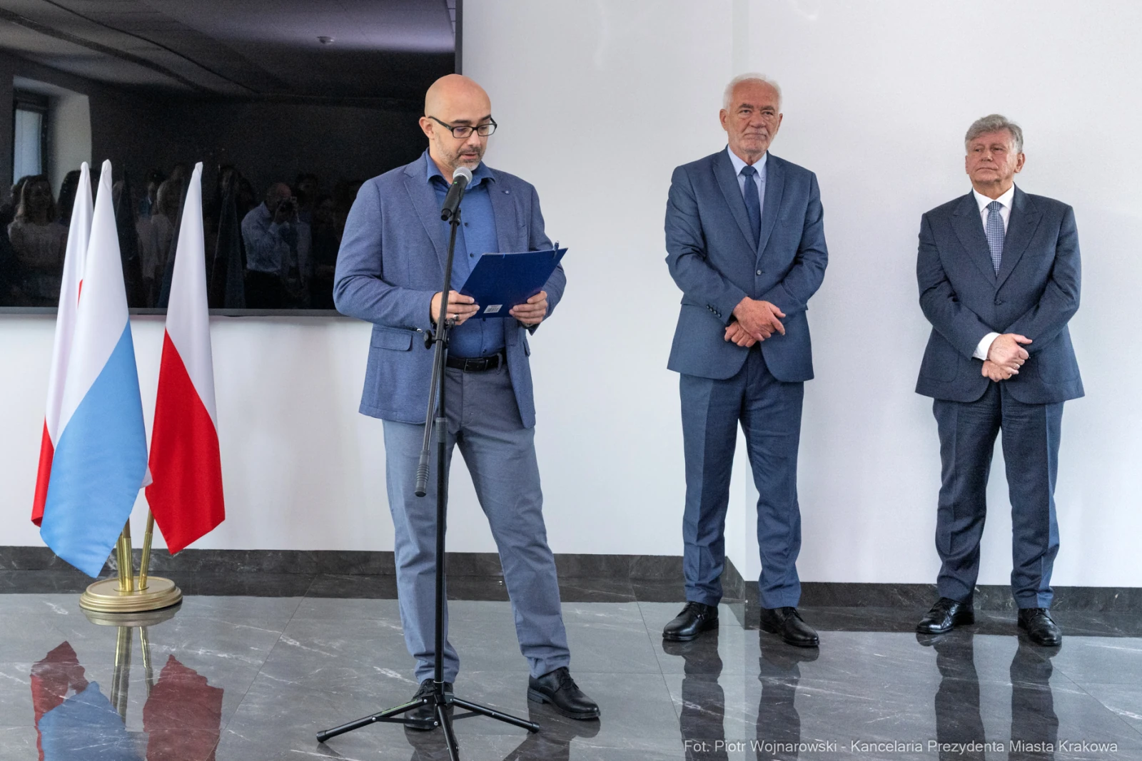 Mężczyzna przemawia przy mikrofonie, obok dwóch innych mężczyzn, w tle flagi.  Autor: Piotr Wojnarowski