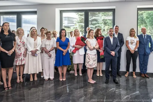 Grupa osób w eleganckich strojach, stojących w sali, niektórzy trzymają kwiaty.