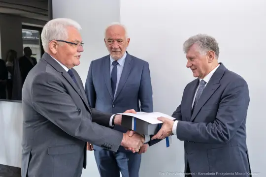 Trzech mężczyzn w garniturach wymienia dokumenty w formalnej atmosferze.