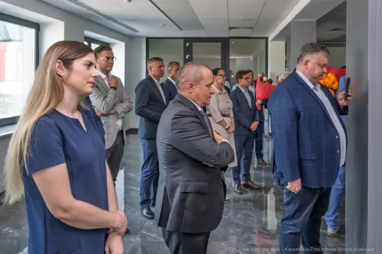 Grupa osób w eleganckich strojach, słuchających przemówienia w nowoczesnym wnętrzu.