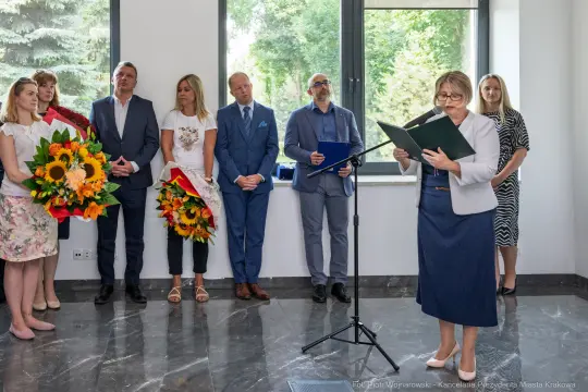 Uroczystość z przemówieniem i wręczeniem kwiatów w eleganckim wnętrzu.