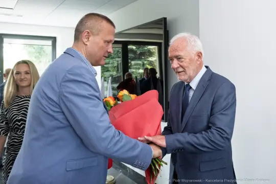 Mężczyzna w garniturze wręcza bukiet kwiatów innemu mężczyźnie w garniturze. W tle kobieta.