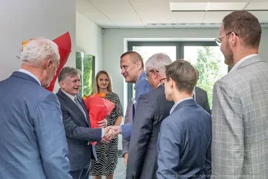 Grupa osób w formalnych strojach, jedna z nich wręcza bukiet kwiatów.