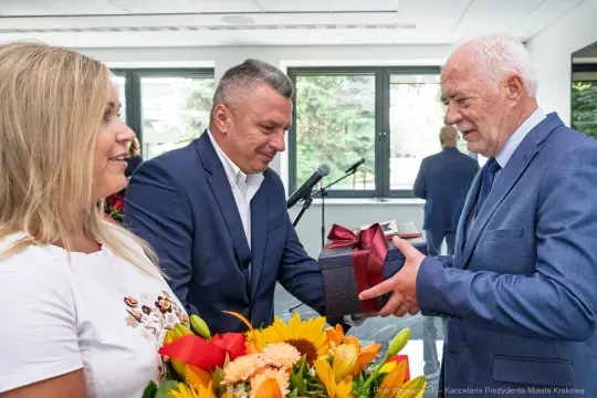 Wręczenie prezentu podczas uroczystości z kwiatami w tle.