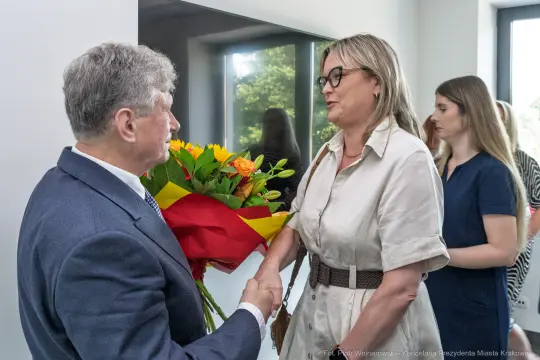 Mężczyzna wręcza bukiet kwiatów kobiecie w eleganckim stroju. W tle widoczna inna kobieta.
