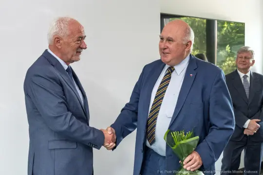Dwaj mężczyźni uścisnęli sobie dłonie, jeden z nich trzyma bukiet kwiatów. W tle stoi kolejny mężczyzna.