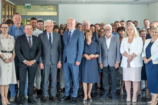 Grupa ludzi w formalnych strojach, uśmiechających się, w biurowym wnętrzu.